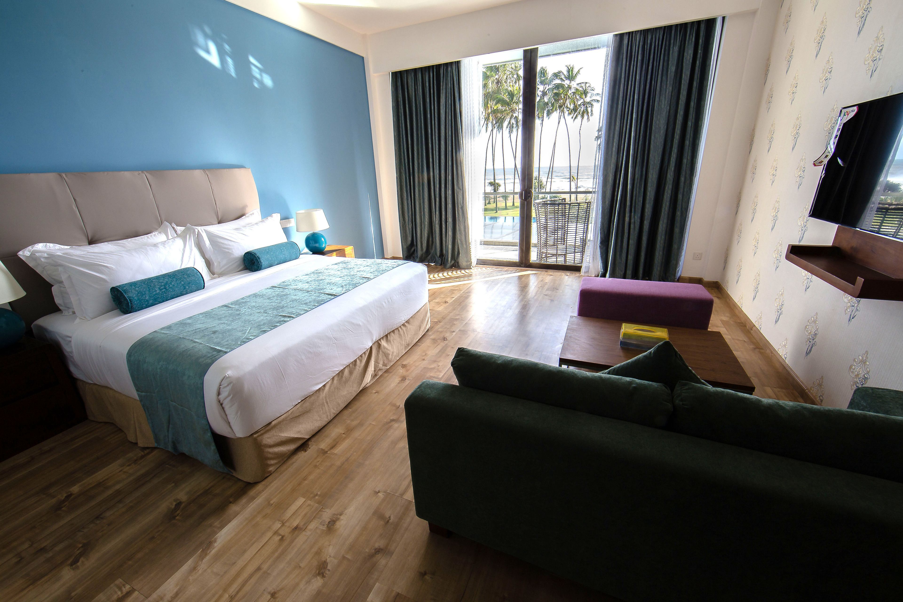 A hotel room with a large bed, a green sofa, a TV, and a balcony with a view of palm trees.