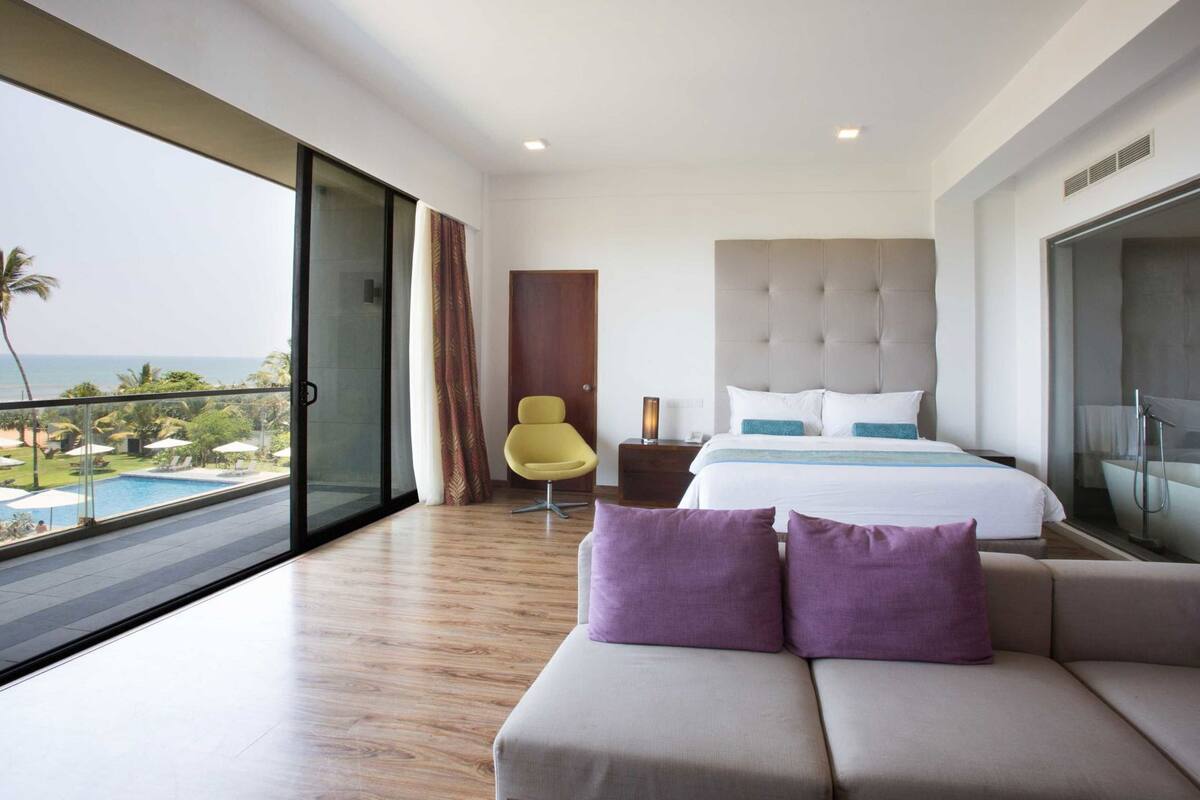 Une chambre d’hôtel moderne, dotée d’un grand lit, d’un canapé, d’un fauteuil, d’une table de chevet et offrant une vue sur l’océan.