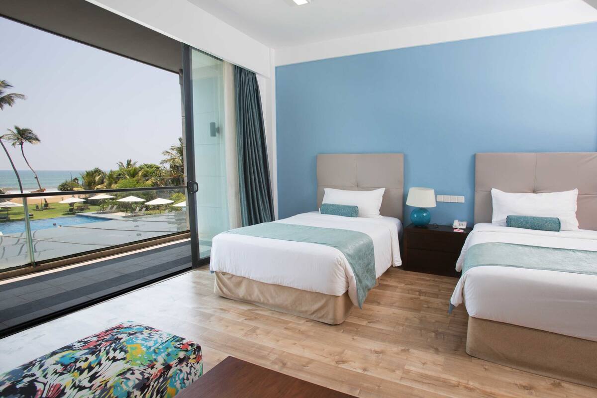 Une chambre d’hôtel avec deux lits, une vue sur une piscine et des palmiers, et un balcon avec une porte vitrée.
