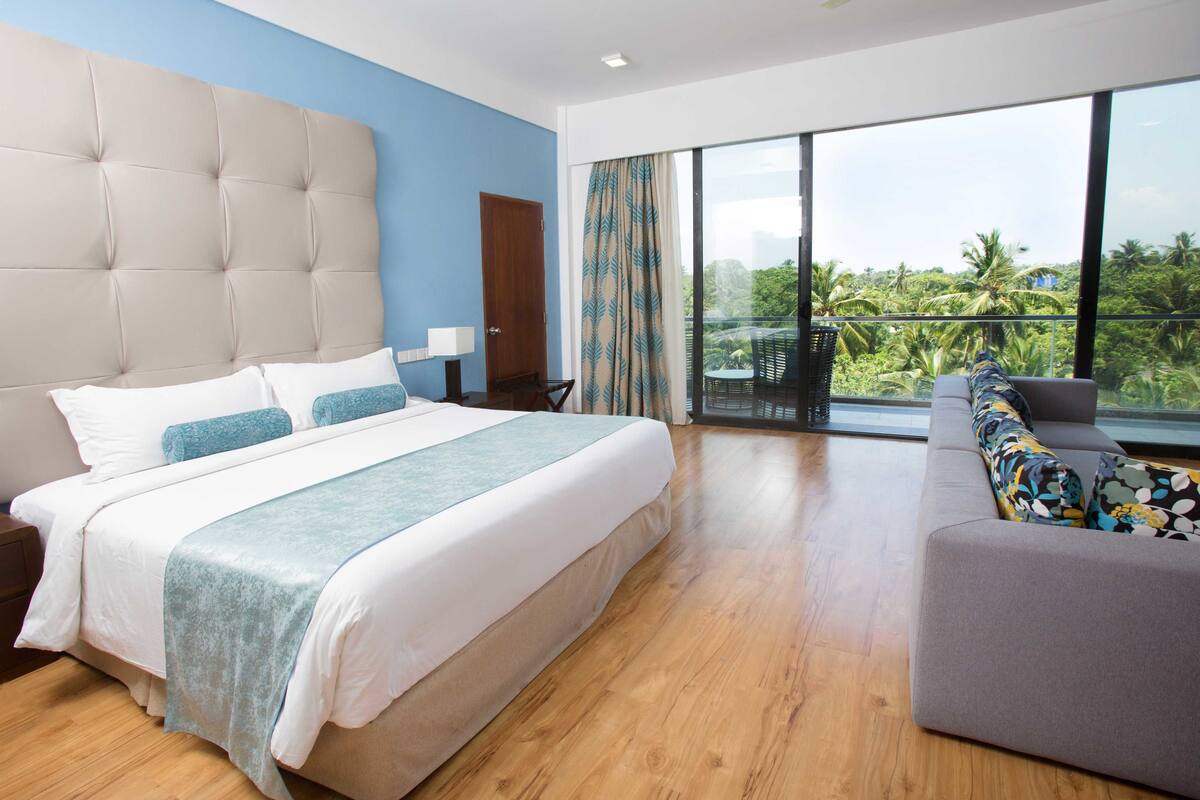 Une chambre d’hôtel moderne avec un grand lit, un canapé et un balcon offrant une vue sur les palmiers.