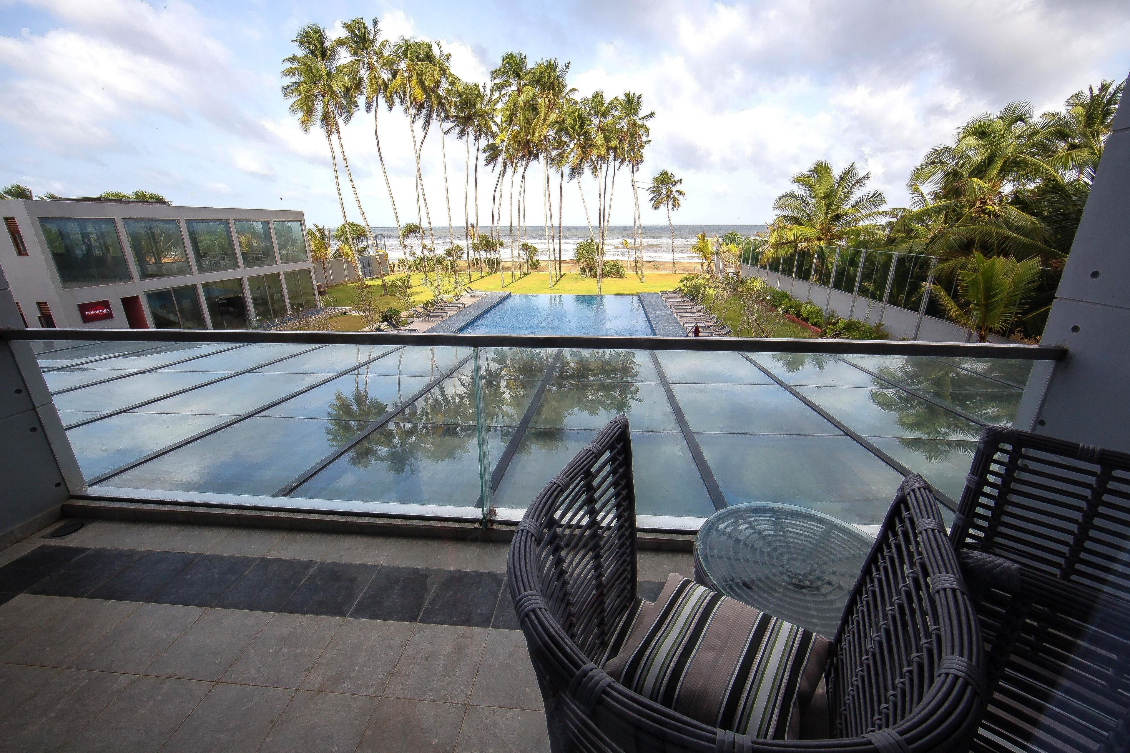 Un balcon doté d’un plancher en verre donnant sur une piscine, des palmiers et une plage.