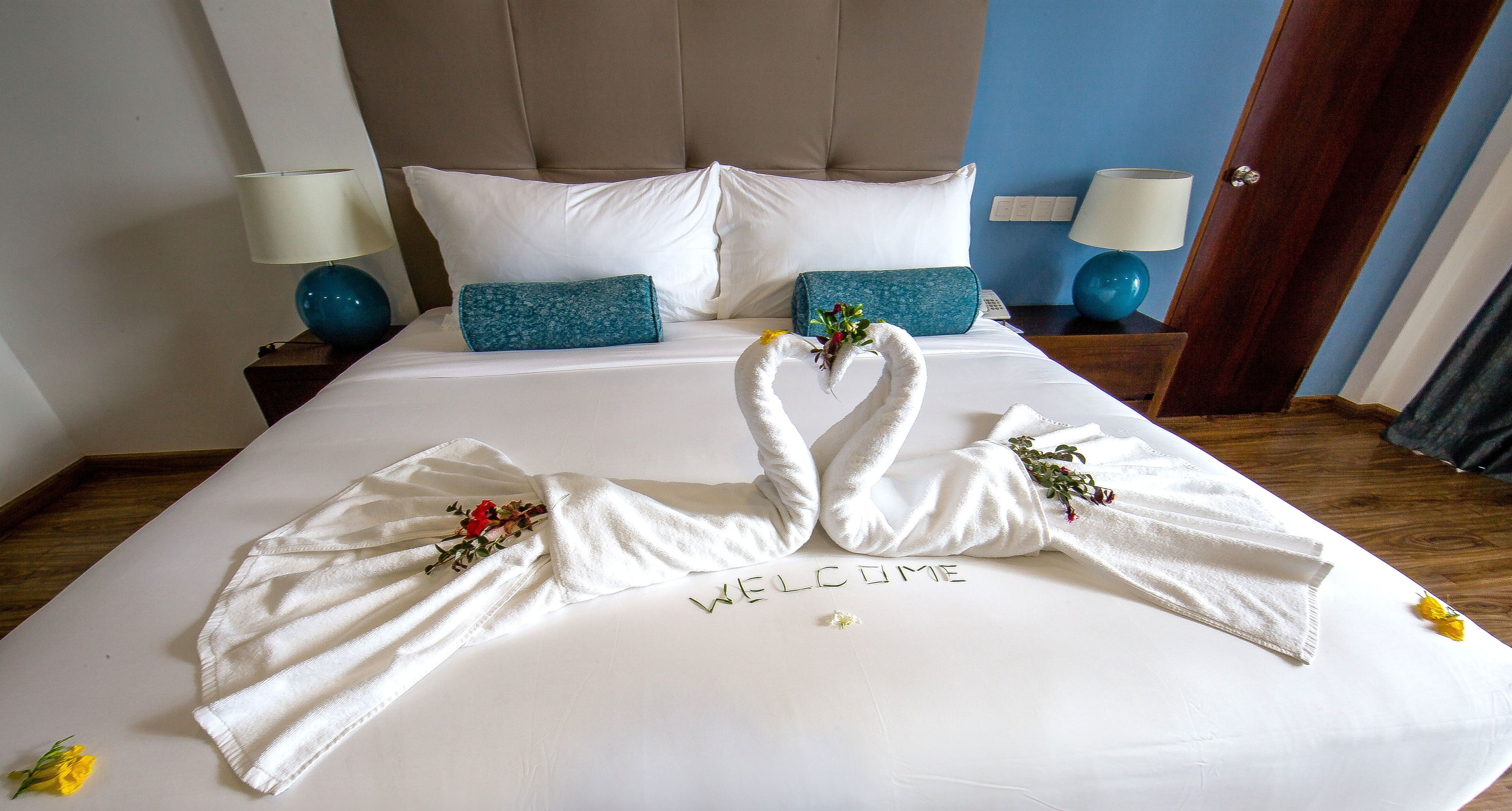 Una cama bien hecha con toallas dispuestas en un gesto acogedor, un cartel que dice “WELCOME” y una flor pequeña sobre la cama.