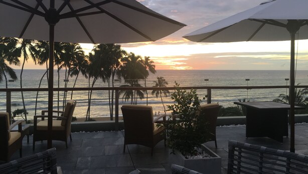 Eine Terrasse mit Sitzgelegenheiten im Freien, Palmen und Blick auf den Ozean bei Sonnenuntergang.