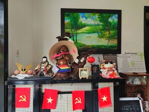 A room with a black wall, a framed painting of a green landscape, a wooden statue with a red hat, a doll, and a sign with shuttle service information. - Flamingo Villa Hoi An (Da Nang)
