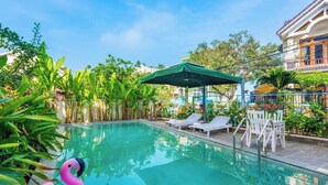 A swimming pool with lounge chairs and umbrellas, surrounded by lush greenery and a building in the background. - Flamingo Villa Hoi An (Da Nang)