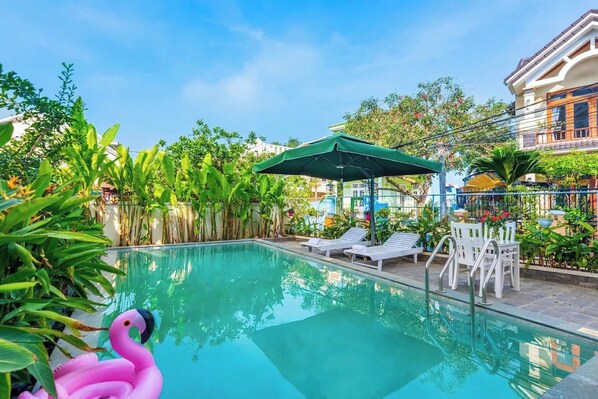 A swimming pool with lounge chairs and umbrellas, surrounded by lush greenery and a building in the background. - Flamingo Villa Hoi An (Da Nang)