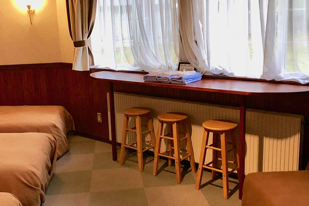 A hotel room with two beds, a window with sheer curtains, and three wooden stools.
