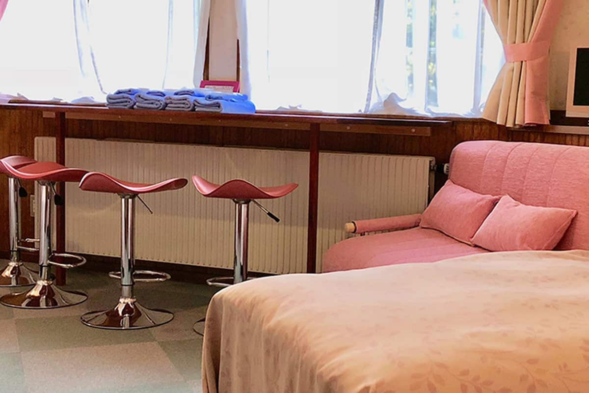 A hotel room with a bed, a TV, a window with curtains, and three bar stools.
