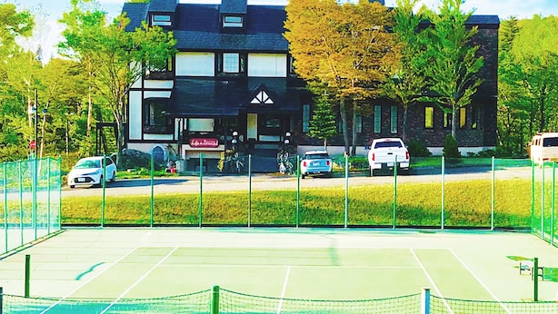Cancha de tenis con red, rodeada por una valla verde. Hay coches aparcados cerca y un edificio al fondo.