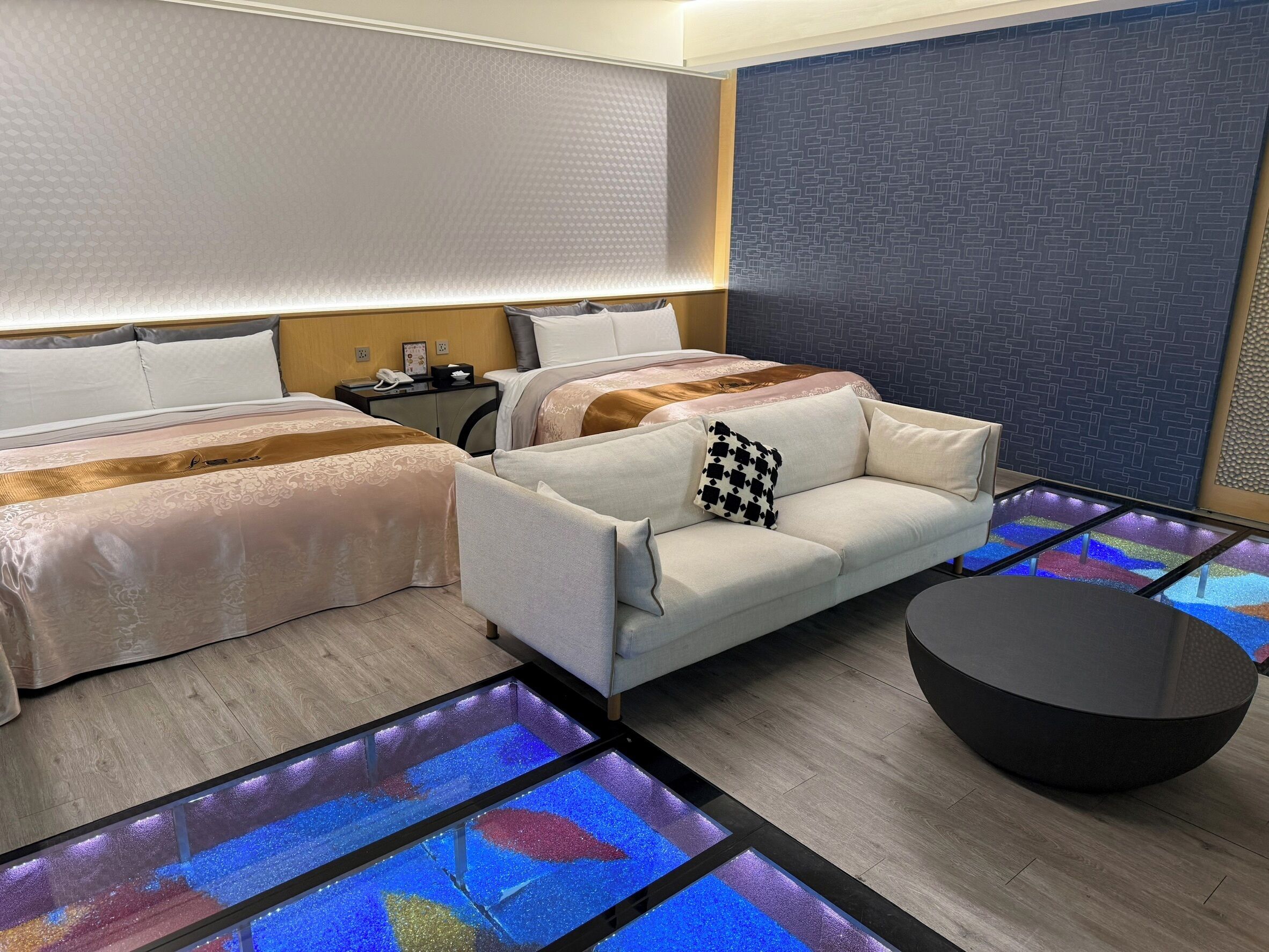 Una habitación de hotel moderna con sofá, dos camas, una mesita de noche y un suelo de vidrio con iluminación de colores.
