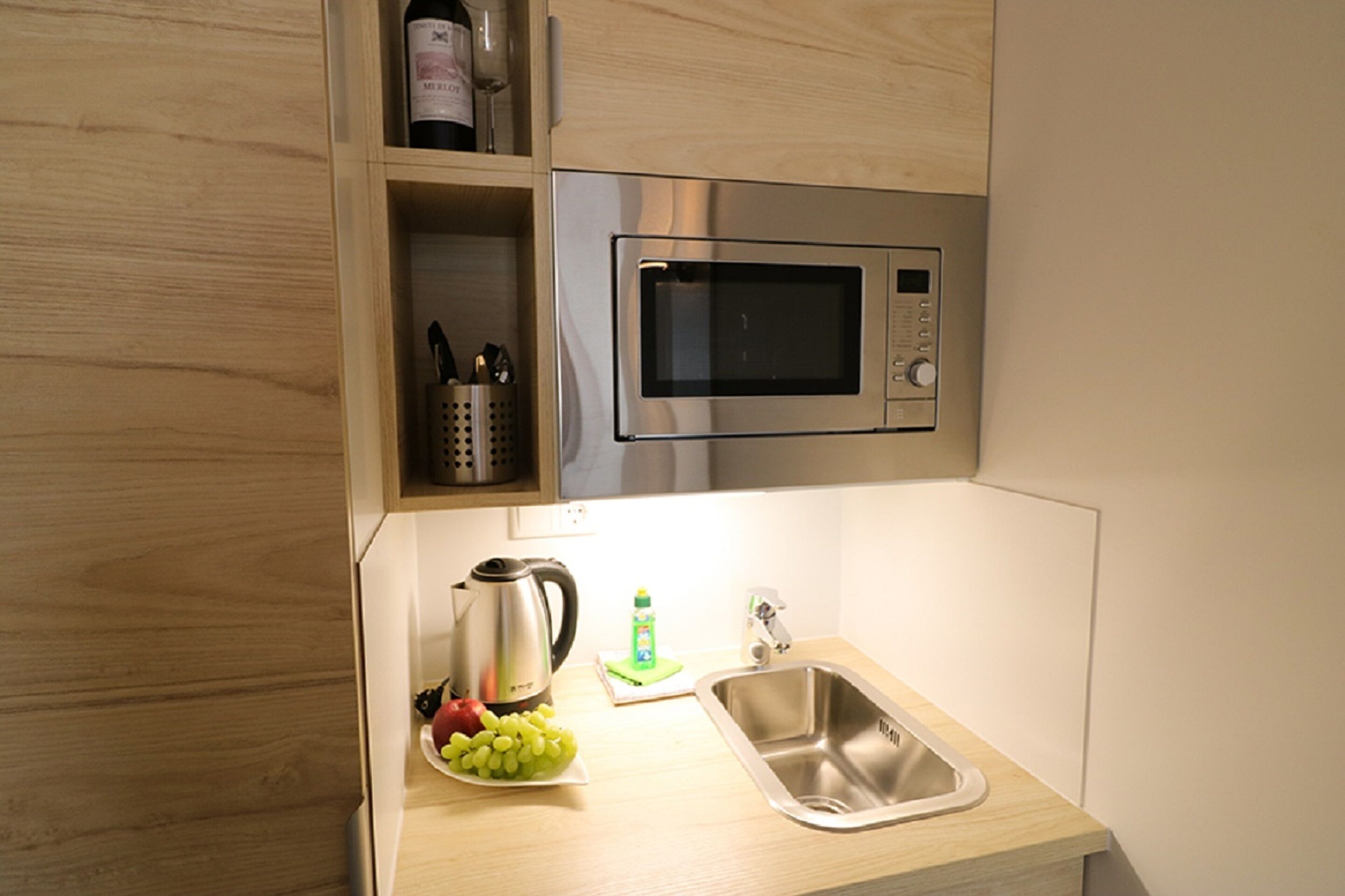 A kitchen with a stainless steel microwave, a sink, a kettle, and a bottle of wine on the counter.