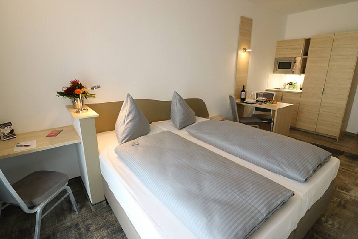 Ein Hotelzimmer mit einem Bett, einem Schreibtisch, einem Sessel, einem kleinen Tisch mit Blumen und einer Küchenzeile.