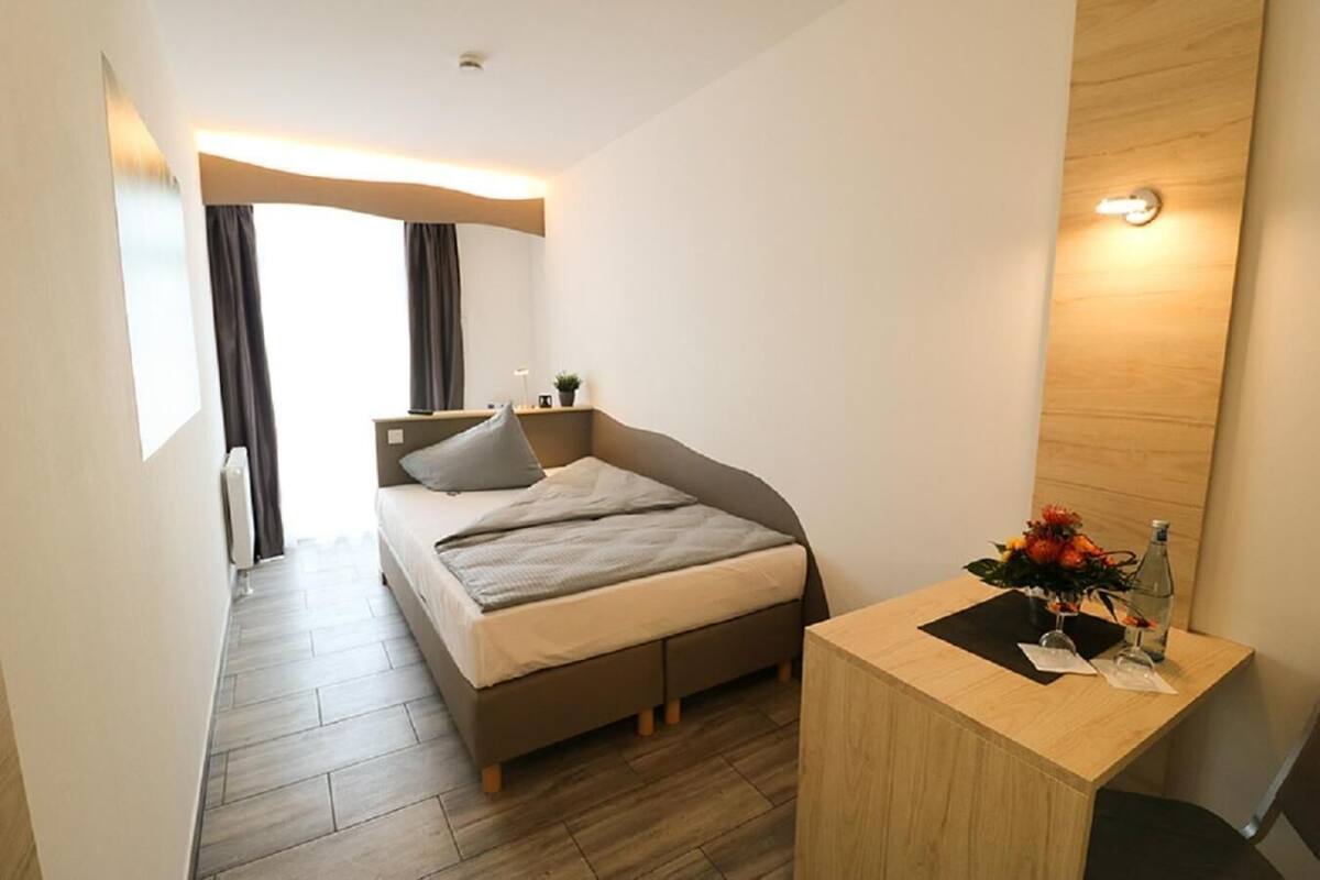 Ein modernes Hotelzimmer mit einem Einzelbett, einem kleinen Holztisch, einem Nachttisch, einer Zimmerpflanze und Blick auf die Stadt.