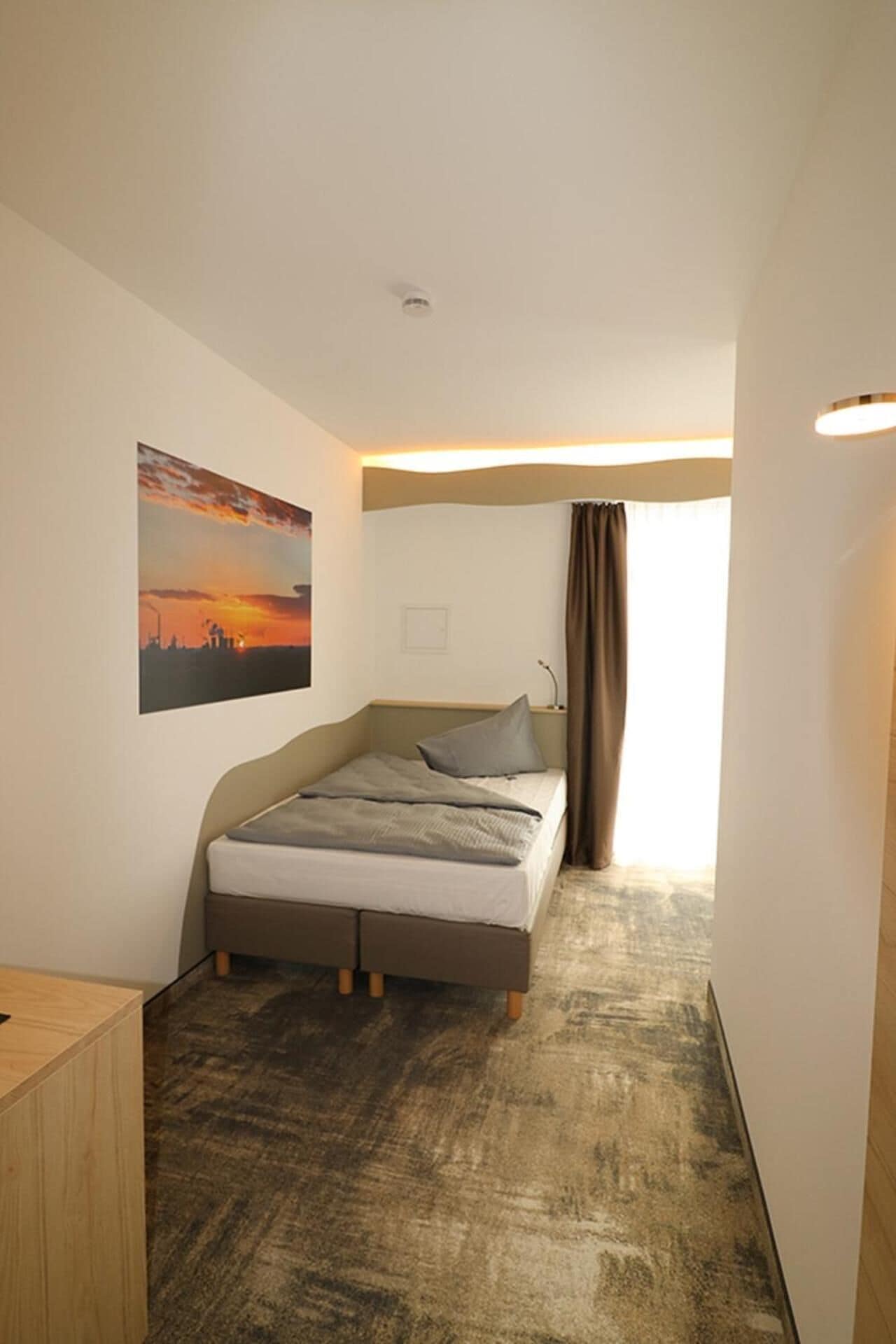 Une petite chambre d’hôtel moderne, avec un lit simple, une table de chevet en bois, un tableau représentant un coucher de soleil et une fenêtre avec des rideaux.