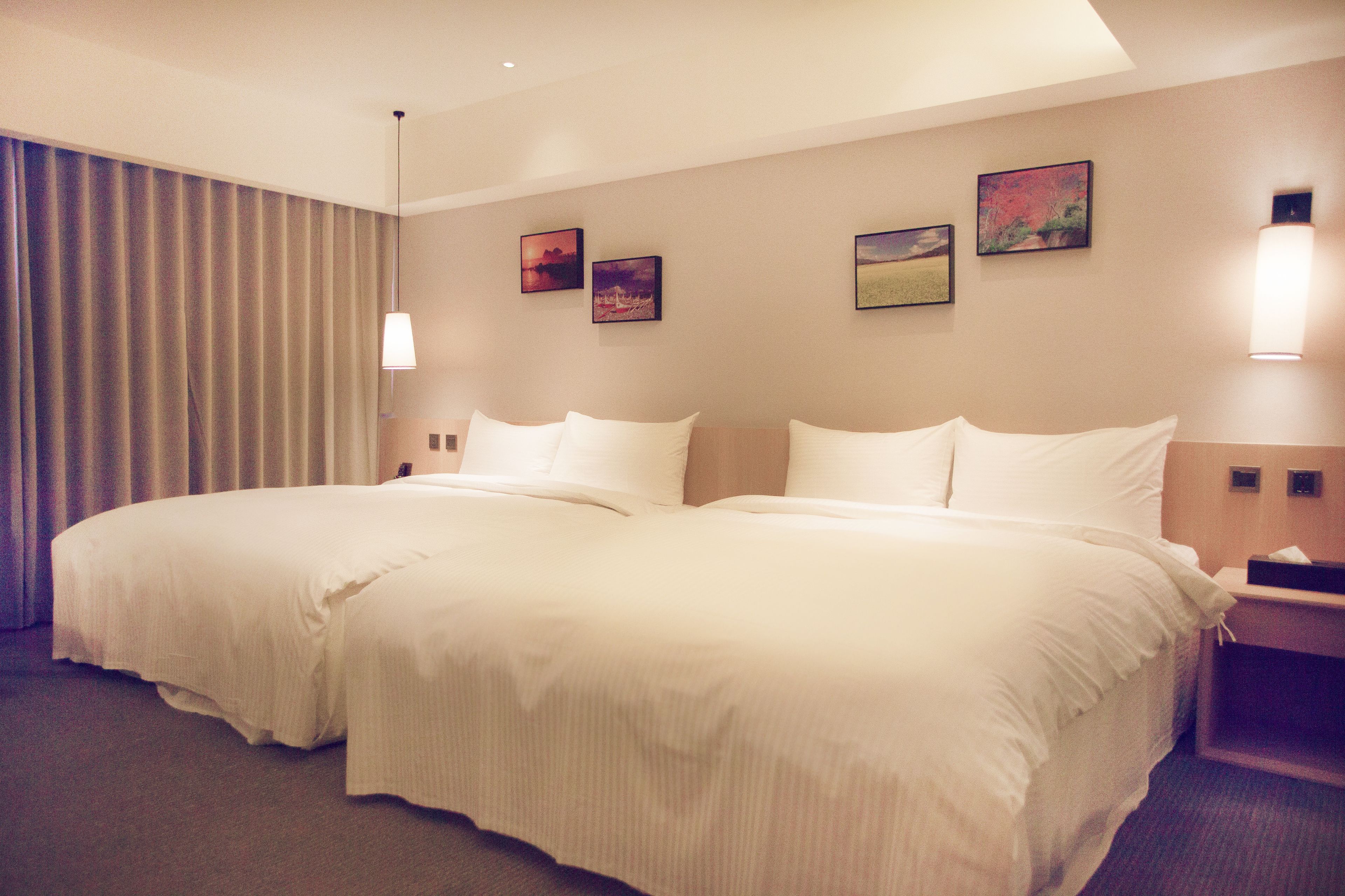 A hotel room with two beds, a large window with curtains, and three wall-mounted paintings.