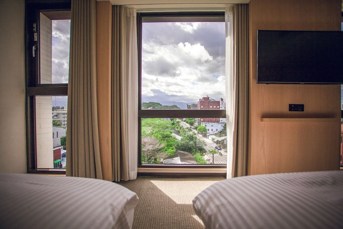 A hotel room with two beds, a large window with curtains, a flat-screen TV mounted on the wall, and a view of the cityscape.