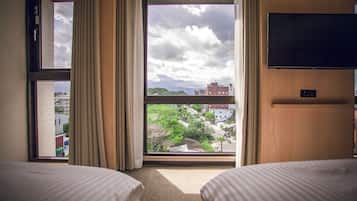 Habitación de hotel con dos camas, un ventanal con cortinas y televisor de pantalla plana montado en la pared, con vista a la ciudad.