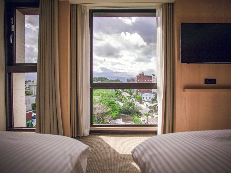 A hotel room with two beds, a large window with curtains, a flat-screen TV mounted on the wall, and a view of the cityscape.