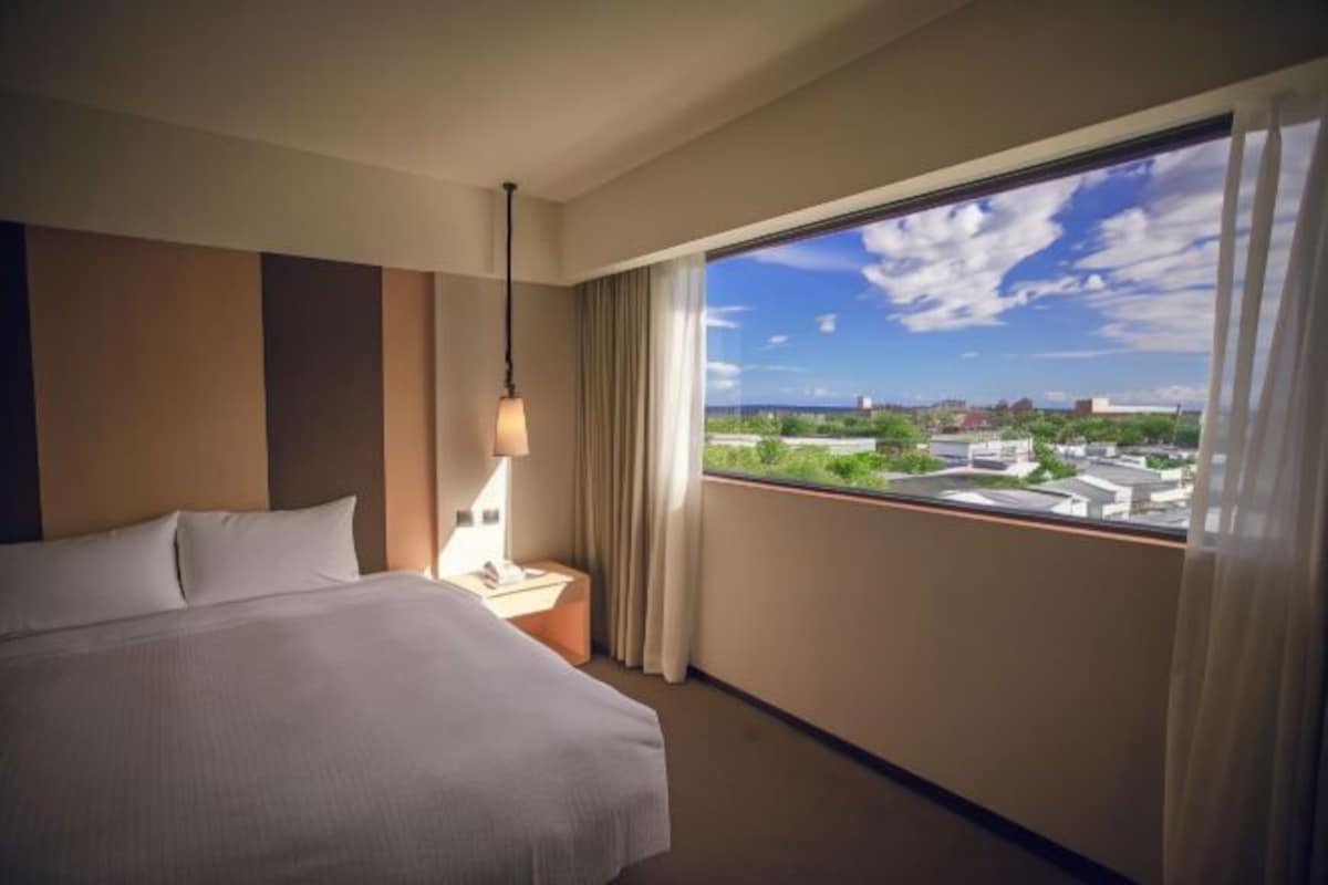 A hotel room with a large bed, a bedside table with a lamp, and a view of a cityscape through a large window.