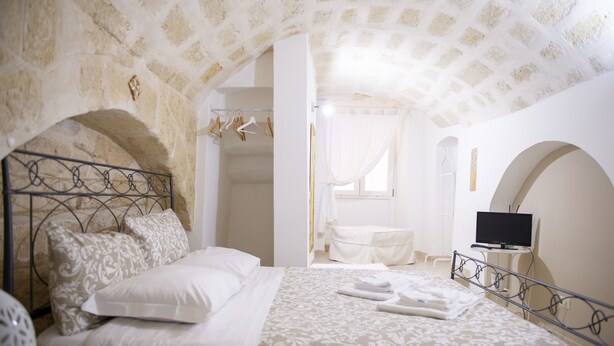 A bedroom with a stone archway, a bed with a metal headboard, a television on a small table, and a window with curtains. - Casa Nave (Monopoli)