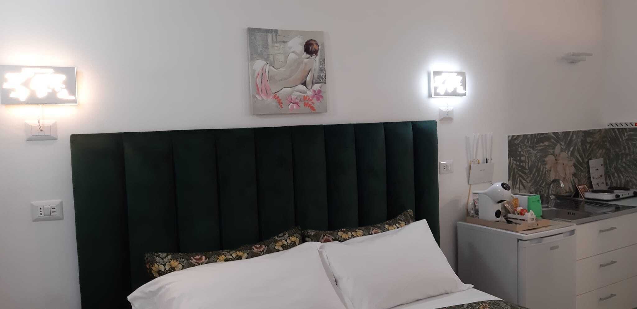 A bedroom with a dark green upholstered headboard, a white bed with floral-patterned pillows, a painting above the bed, and a small kitchenette with a sink and a hairdryer on the counter.