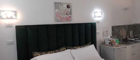 A bedroom with a dark green upholstered headboard, a white bed with floral-patterned pillows, a painting above the bed, and a small kitchenette with a sink and a hairdryer on the counter.