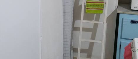 A narrow bathroom with a white sink, a white toilet, a white shelf with a red towel, a white ladder, and a white curtain.