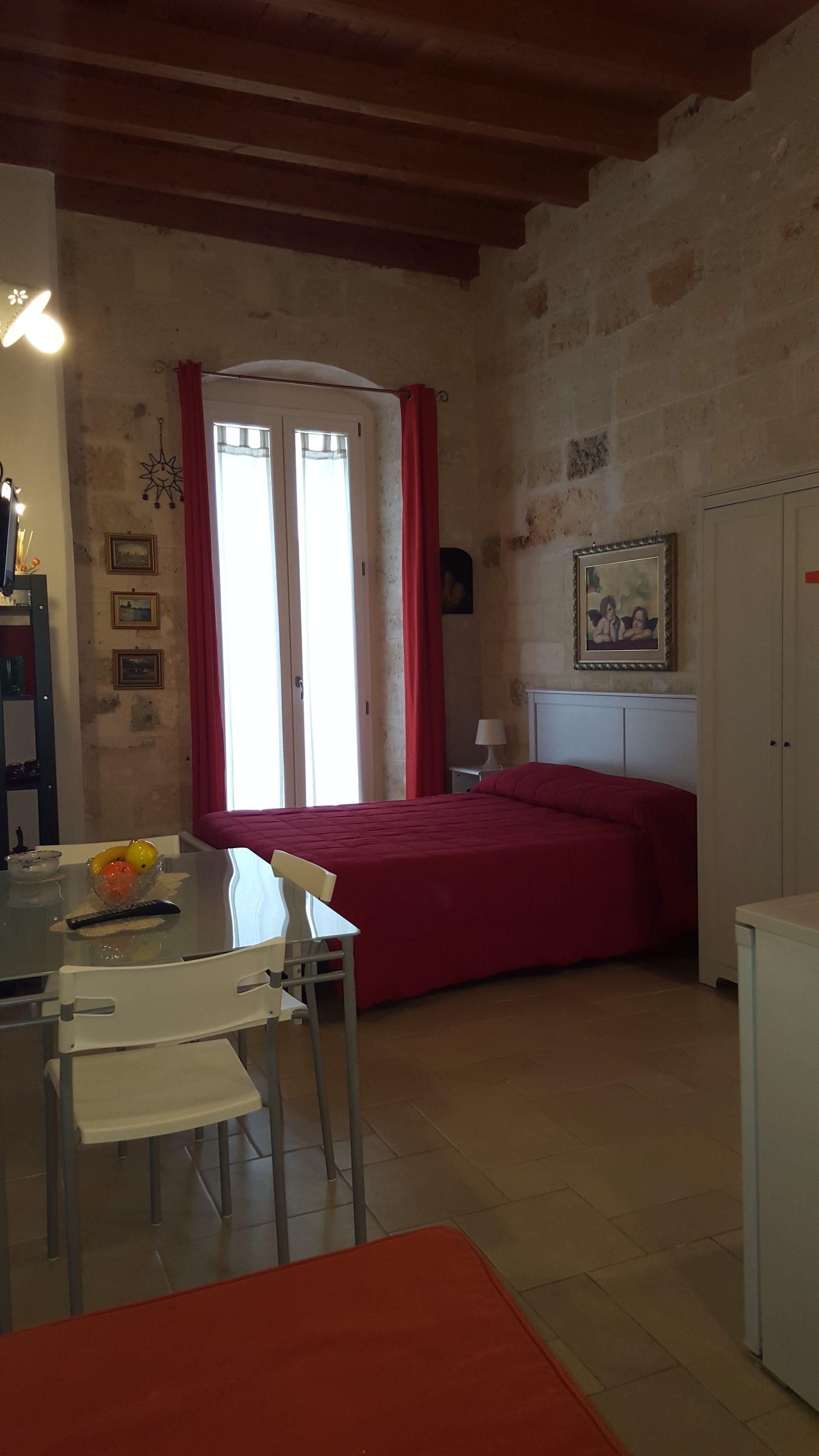 Une pièce comprenant un lit, une table avec des chaises, une petite table sur laquelle se trouve un saladier rempli de fruits, une lampe et un tableau accroché au mur.