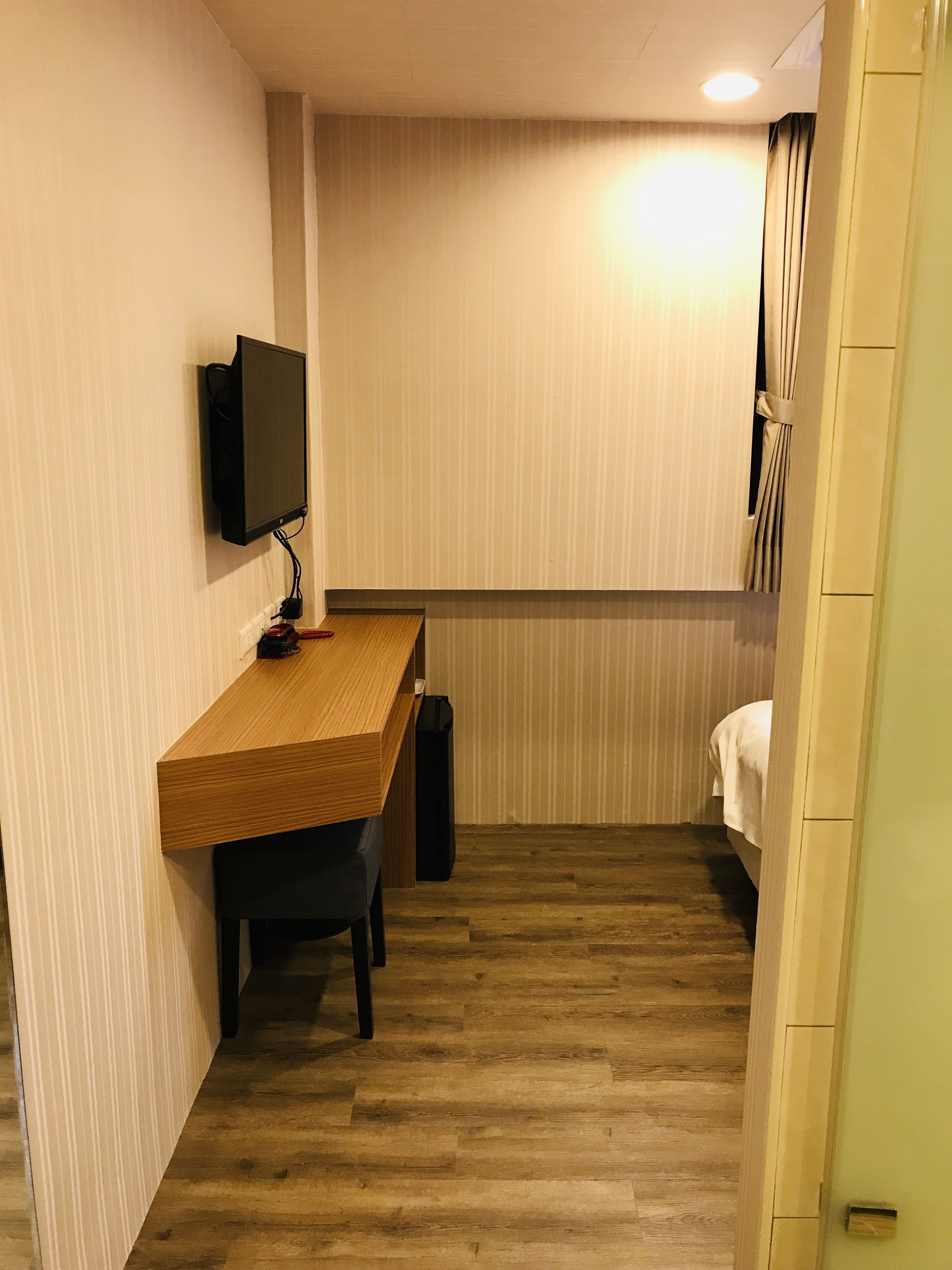 A hotel room with a wooden desk, a flat-screen TV mounted on the wall, a black suitcase, and a bed with white bedding.