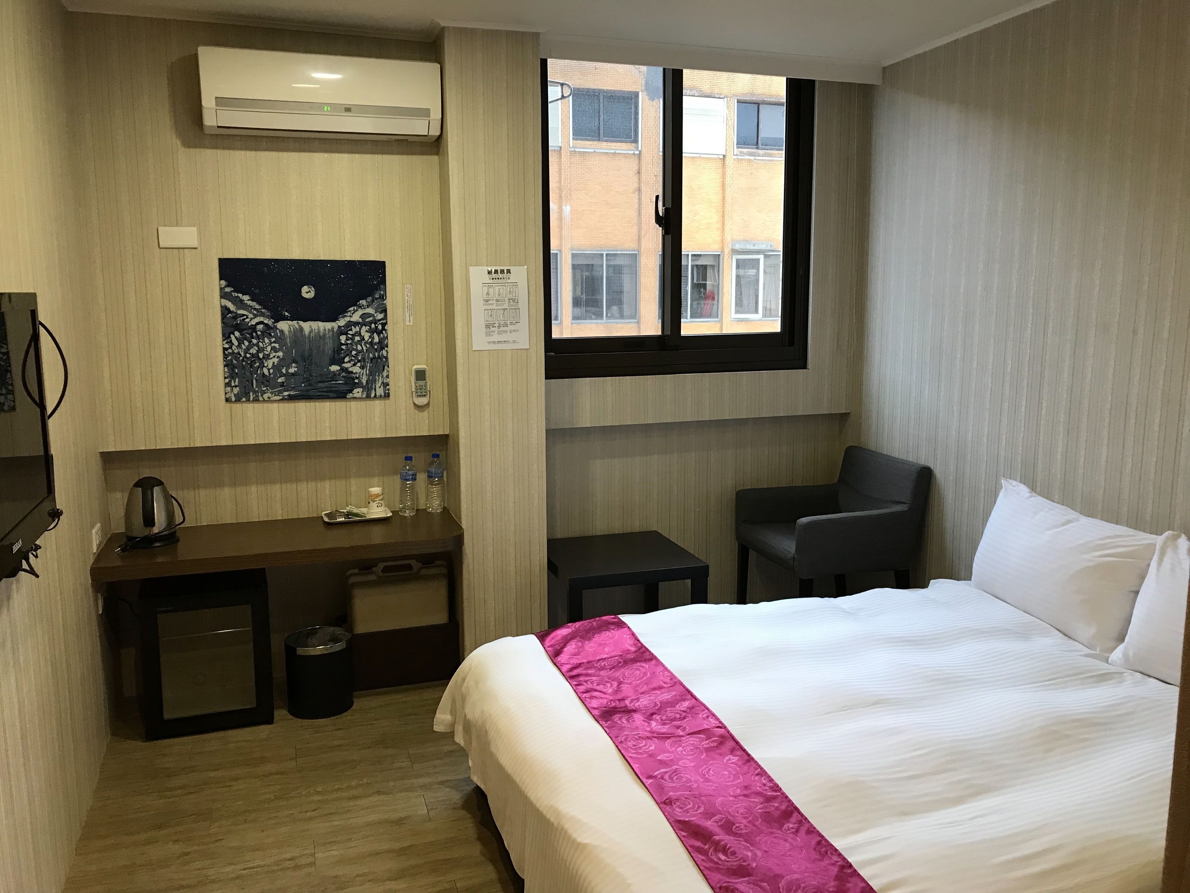A hotel room with a bed, a desk, a chair, an air conditioner, a window, and a painting on the wall.