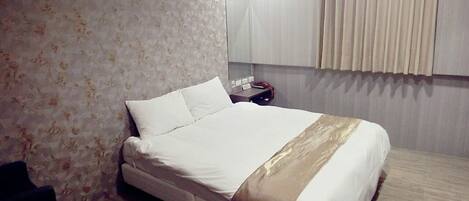 Una cama bien tendida con colcha blanca y color crema, una mesita de noche con lámpara y una pared cubierta de papel tapiz floral.