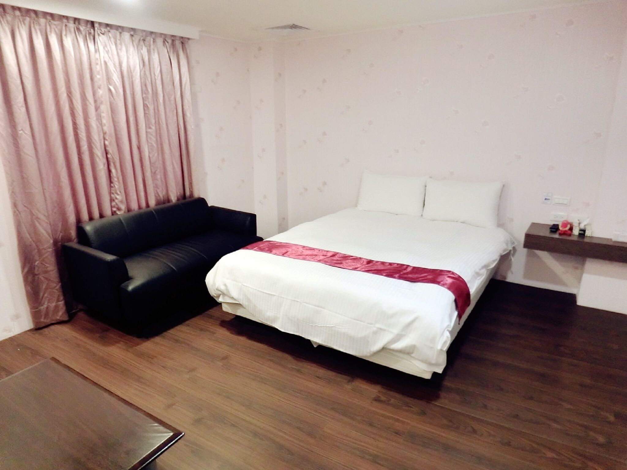 A bedroom with a bed, a black sofa, a wooden floor, and a window with curtains.