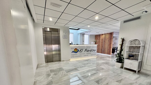 A modern reception area with a marble floor, a reception desk, an elevator, and a decorative plant. - Hotel El Paraiso (Aguilas)
