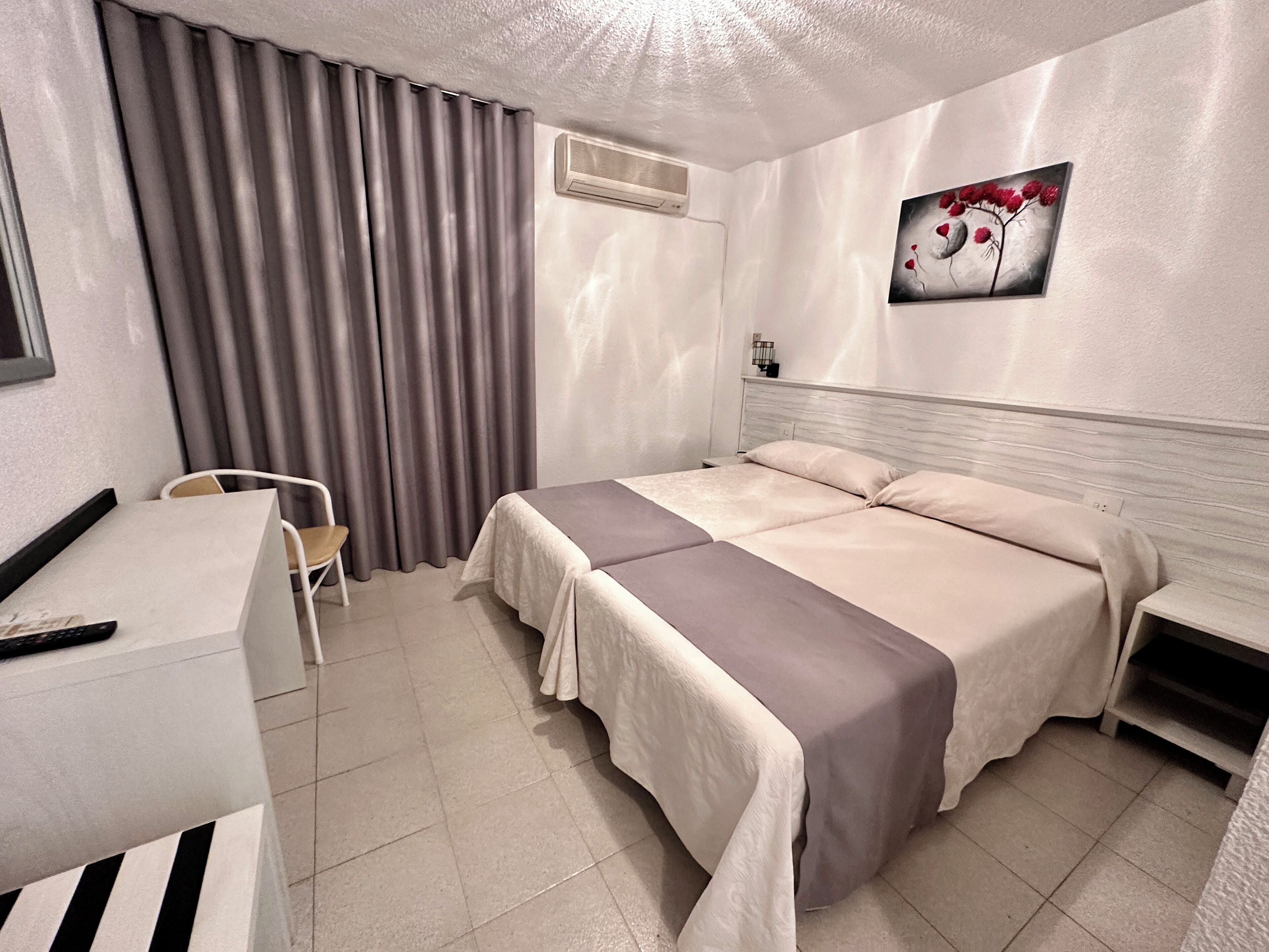 A hotel room with a bed, a desk, a chair, a painting on the wall, and a window with curtains.