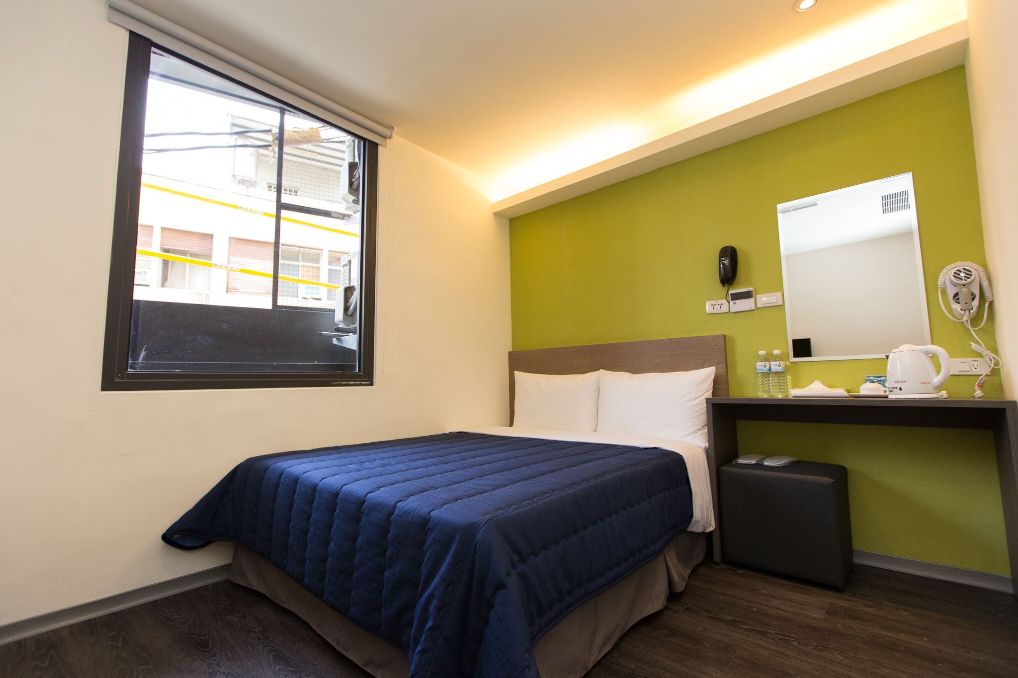 A hotel room with a bed, a small desk with a mirror, a telephone, and a window with a view of buildings.