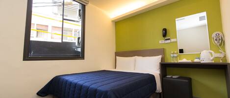 A hotel room with a bed, a small desk with a mirror, a telephone, and a window with a view of buildings.