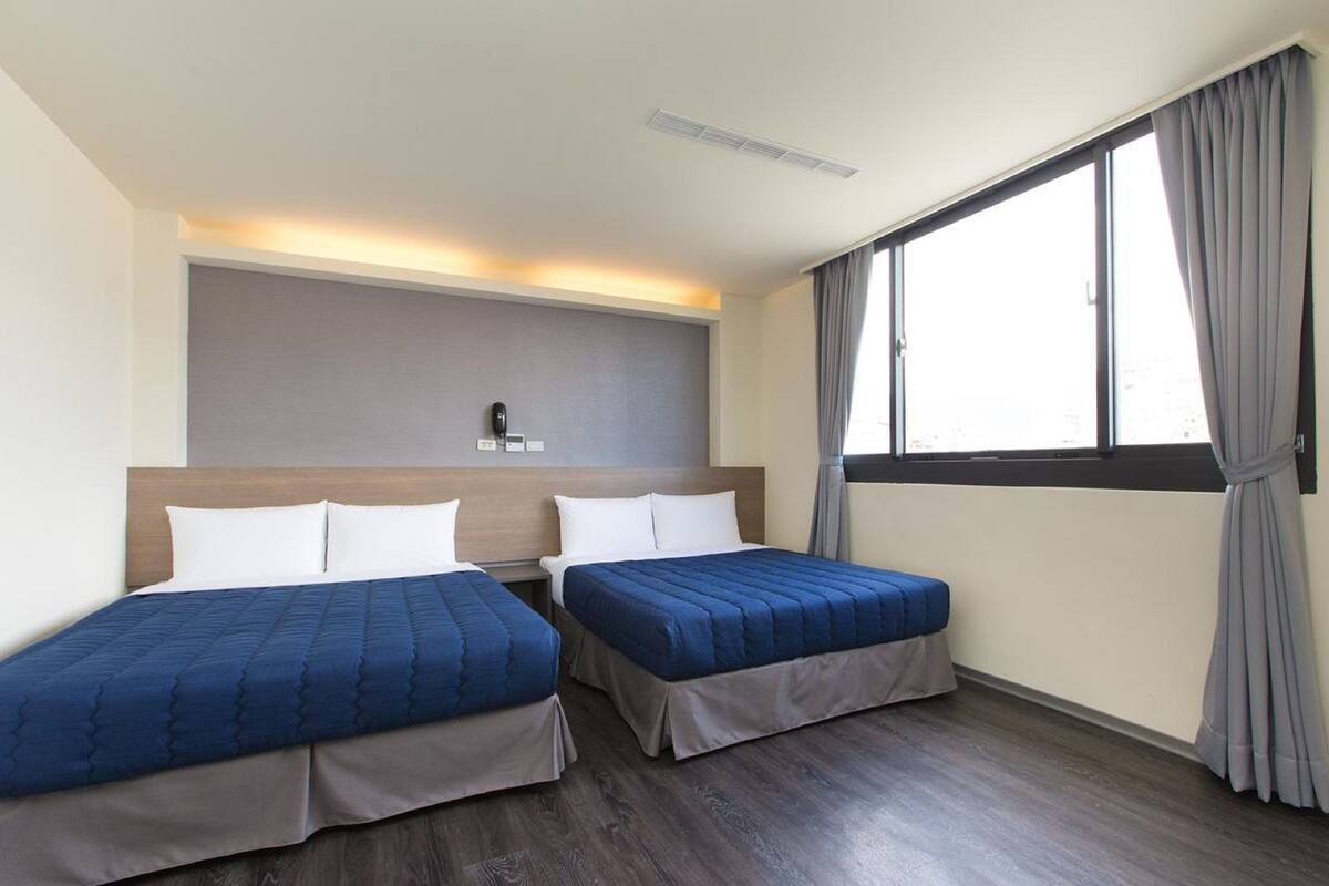 A hotel room with two beds, a large window with curtains, a headboard with a phone, and a wall-mounted light.