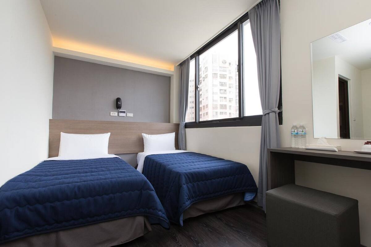 A hotel room with two beds, a desk, a chair, a large window, and a mirror.