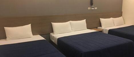 Quarto de hotel com três camas, cada uma com almofadas brancas e colchas azul-marinho. Há um telefone fixado na parede e duas tomadas elétricas.