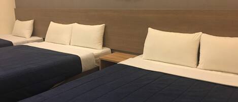 Habitación de hotel con dos camas, cada una con ropa blanca y cabeceros azul oscuro. Hay una mesita de noche entre las camas, y se observan un teléfono y un termostato fijados a la pared.