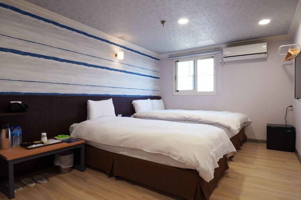A hotel room with two beds, a small table with a water bottle, a trash bin, and an air conditioning unit.