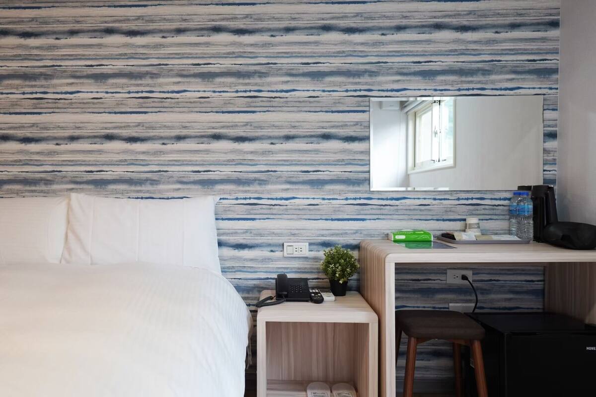 A hotel room with a bed, a desk with a computer, a phone, and a small plant.
