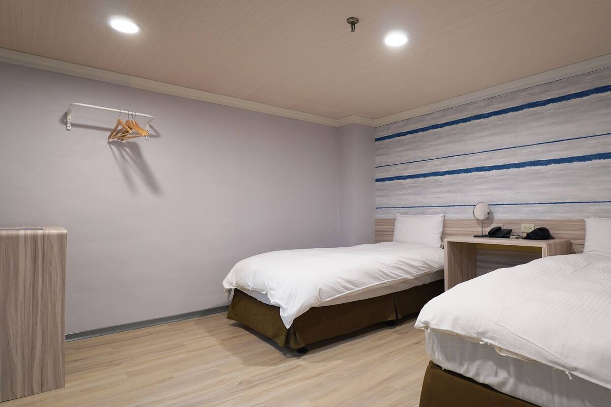 A hotel room with two beds, a bedside table with a lamp, a hanging clothes rack, and a wall with horizontal stripes.
