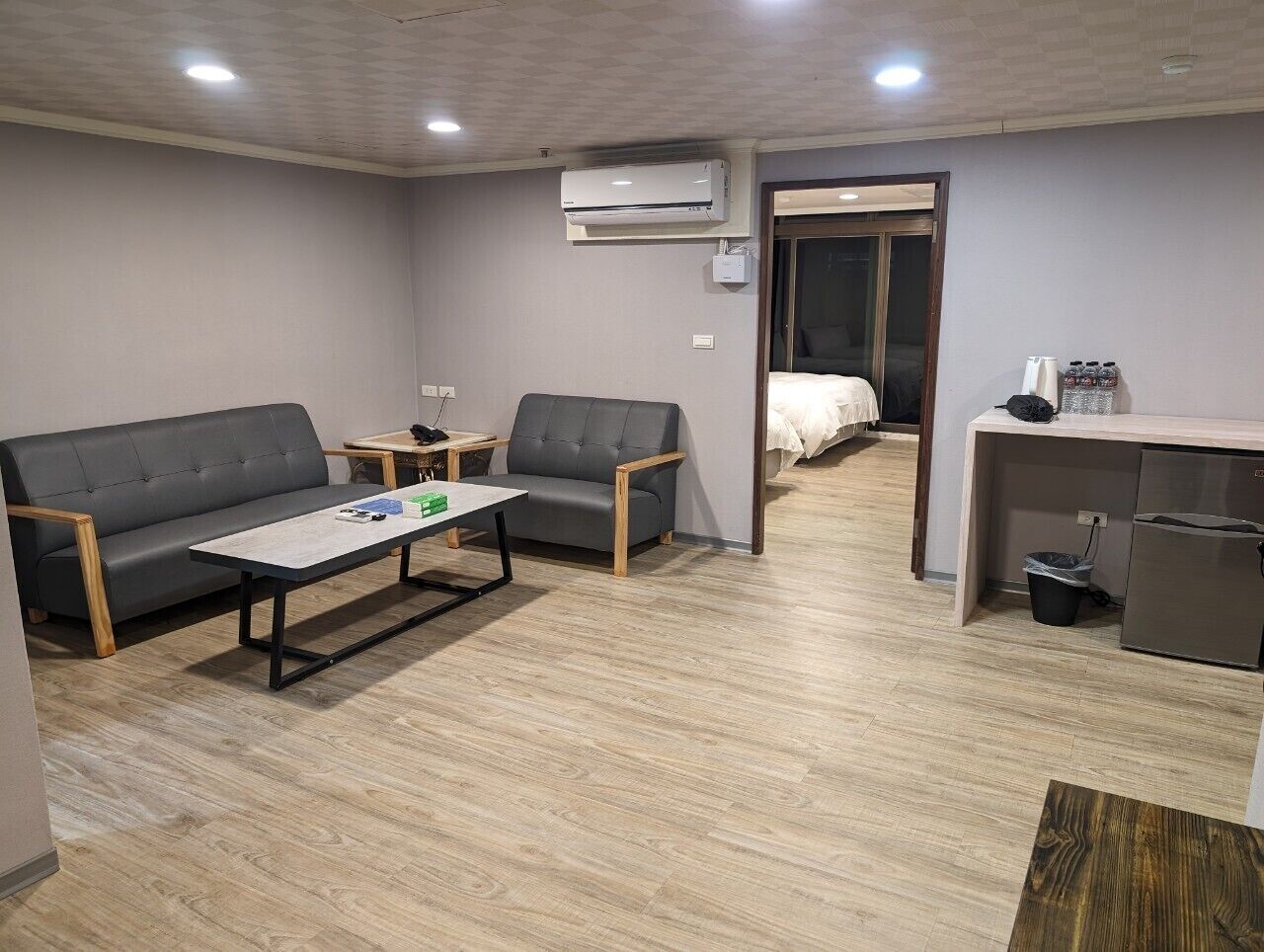 A room with a sofa, a chair, a small table, a kitchenette, and a bed in the background.