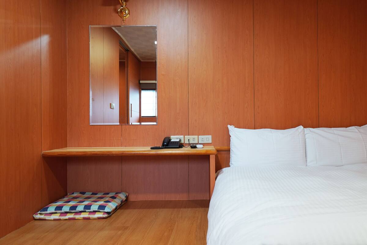 A hotel room with a wooden wall, a bed with white bedding, a bedside table with a phone and a lamp, and a colorful cushion on the floor.
