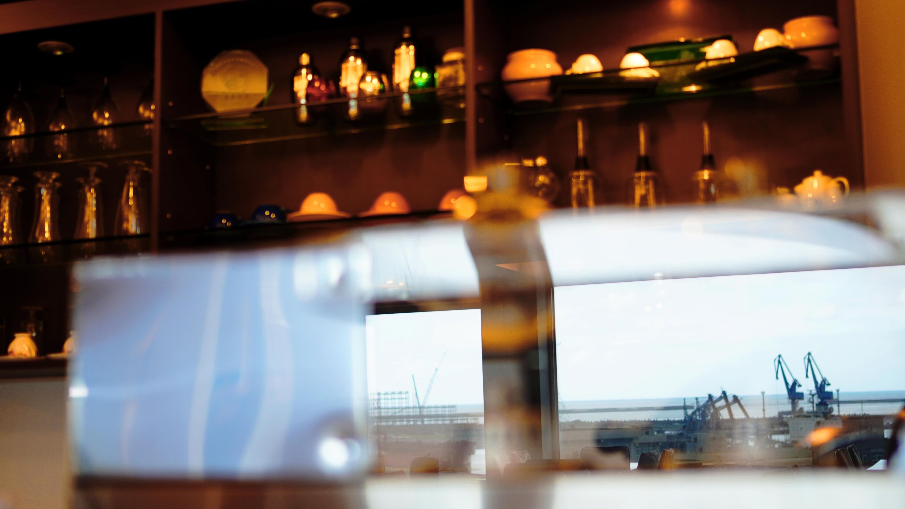 A bar area with shelves of bottles and glasses, a large window with a view of a port and cranes.