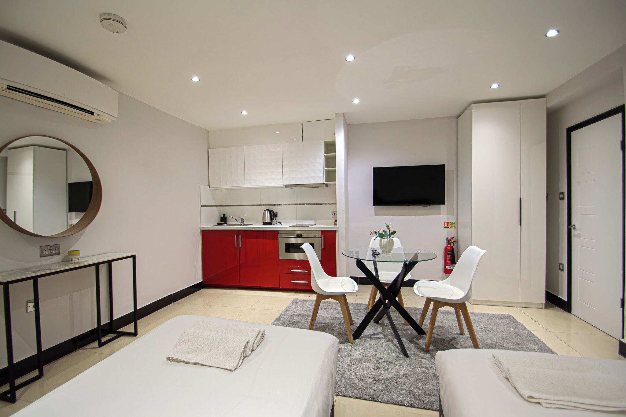 A modern hotel room with a bed, two white chairs, a small table, a TV, a kitchen area, and a mirror.