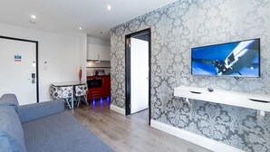 A modern living room with a grey sofa, a TV mounted on the wall, and a kitchen in the background. - Imperial Court Suites (London)