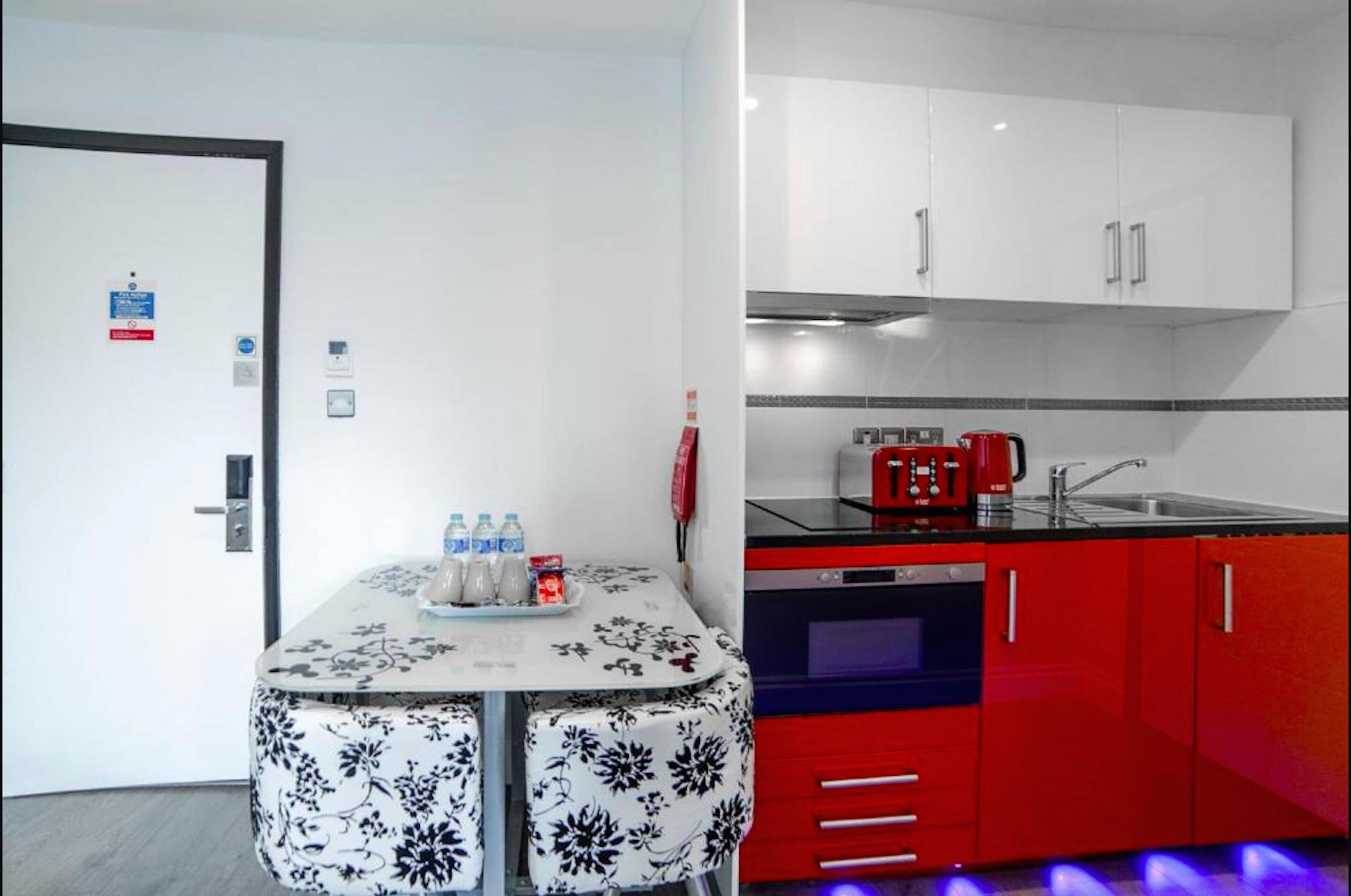 A room with a white door, a table with a floral tablecloth, and a red kitchen with cabinets and appliances.