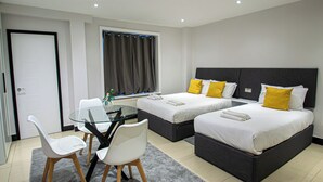 A hotel room with two beds, a small table with chairs, a window with curtains, and a door. - Imperial Court Suites (London)